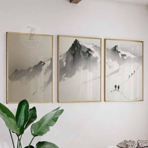 Peut inclure: Trois impressions encadrées de paysages de montagne. L'œuvre présente un style minimaliste avec des représentations de montagnes enneigées en gris et blanc, semblables à des aquarelles. Les cadres sont dorés et les impressions sont exposées sur un mur blanc.