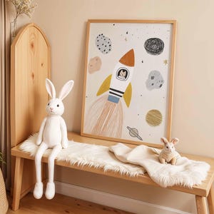 May include: A wooden bench with a white faux fur throw, a large white bunny plush, and a framed space-themed art print. The art features a rocket ship with a cat in the window, planets, and stars. A small bunny plush sits on the throw.