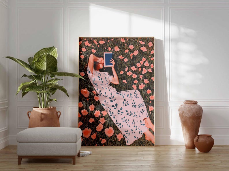 K&ouml;nnte beinhalten: Ein gro&szlig;er gerahmter Kunstdruck zeigt eine Person, die in einem Blumenfeld liegt und ein Tablet h&auml;lt. Die Person tr&auml;gt ein rosa Kleid mit floralem Muster. Das Kunstwerk wird in einem Raum mit einer Pflanze, einem Ottoman und dekorativen Vasen ausgestellt.