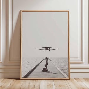 Puede incluir: Impresi&oacute;n en blanco y negro enmarcada que muestra a una mujer en un muelle de madera con un avi&oacute;n acerc&aacute;ndose. El mar y el cielo crean un fondo sereno. La impresi&oacute;n est&aacute; en un marco de madera.