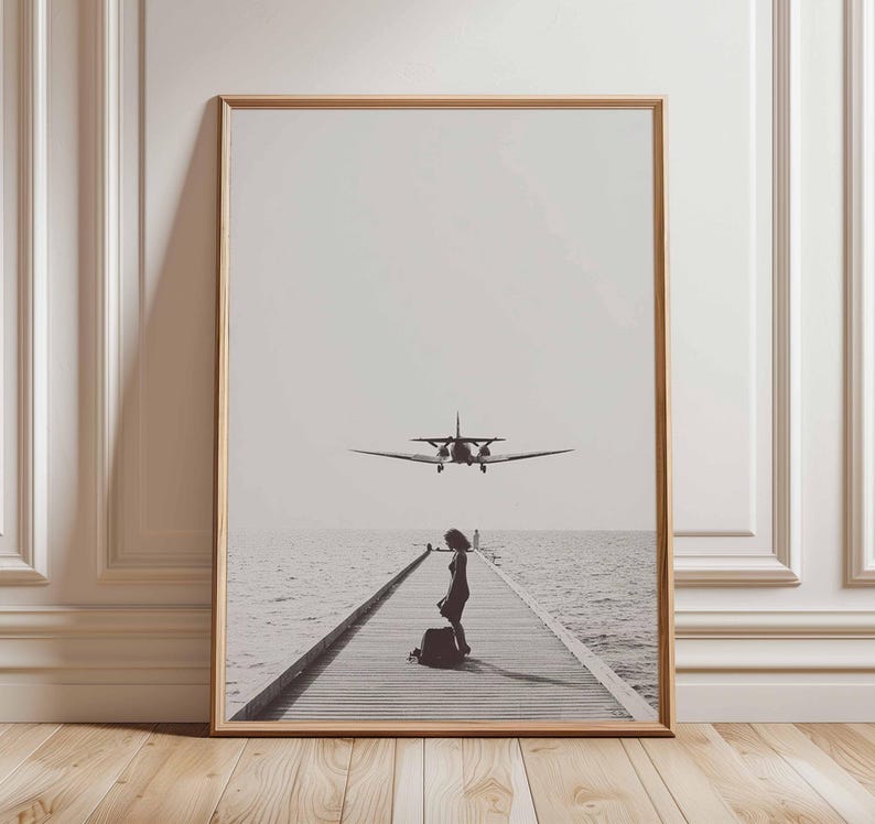 Puede incluir: Impresi&oacute;n en blanco y negro enmarcada que muestra a una mujer en un muelle de madera con un avi&oacute;n acerc&aacute;ndose. El mar y el cielo crean un fondo sereno. La impresi&oacute;n est&aacute; en un marco de madera.