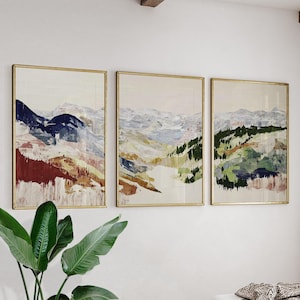 May include: Three framed abstract landscape paintings. Each features a mountain scene with a palette of blues, greens, and earth tones. The frames are gold, and the artwork is displayed on a white wall.