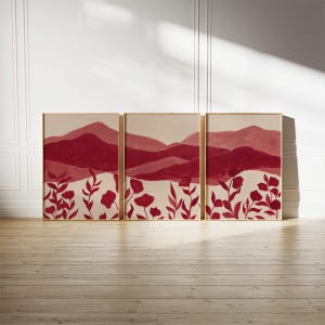 May include: Triptych of framed art prints. Each print features a landscape of burgundy mountains and red floral elements against a cream background. The artwork is displayed on a light wood floor against a white wall.