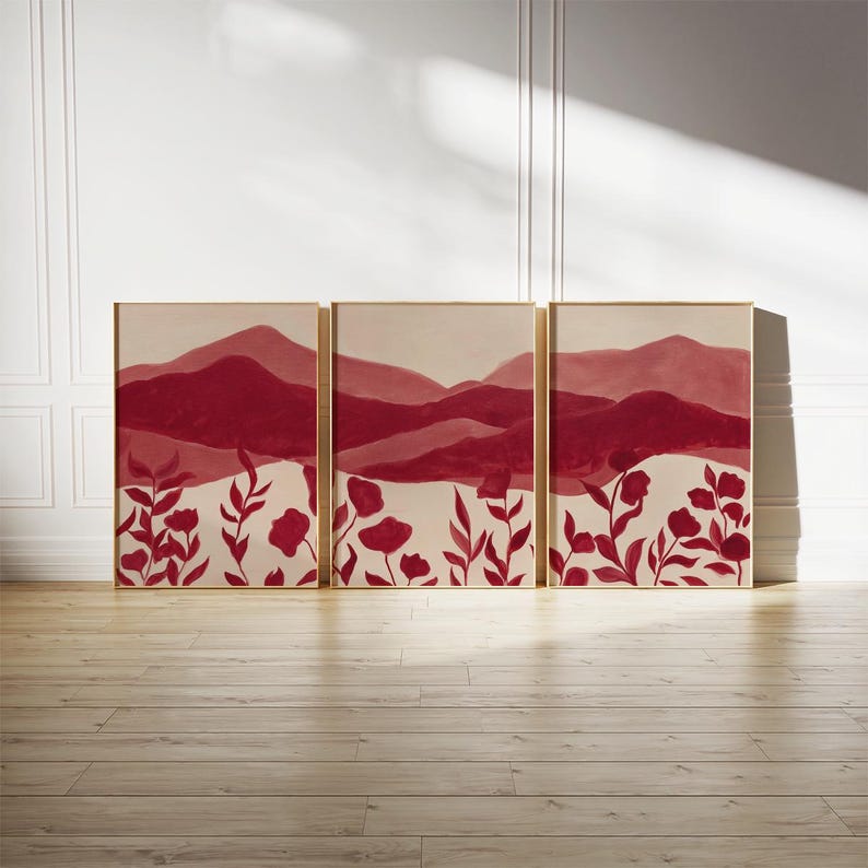 May include: Triptych of framed art prints. Each print features a landscape of burgundy mountains and red floral elements against a cream background. The artwork is displayed on a light wood floor against a white wall.