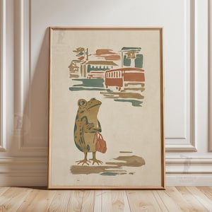 May include: Framed vintage-style print featuring a frog with a bag, standing in front of a bus stop scene. The artwork uses muted tones of brown, green, and orange. The print is in a wooden frame and is displayed on a neutral wall.