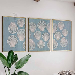 May include: Three framed artworks with white seashell patterns on a pale blue backdrop. The frames have a gold finish. The art is displayed on a white wall, with a green plant in the foreground.