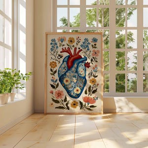 May include: A framed art print featuring a stylized anatomical heart illustration. The heart is depicted in shades of blue and red, adorned with floral patterns and surrounded by colorful flowers and foliage. The artwork is displayed in a bright room with natural light.
