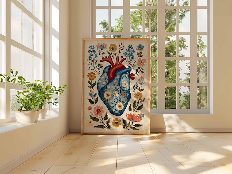 May include: A framed art print featuring a stylized anatomical heart illustration. The heart is depicted in shades of blue and red, adorned with floral patterns and surrounded by colorful flowers and foliage. The artwork is displayed in a bright room with natural light.