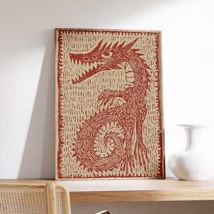 May include: Framed print featuring a red dragon illustration on a beige background. The dragon has sharp teeth and claws, with a coiled tail. The artwork is in a wooden frame and rests on a wooden shelf. A white vase is next to the print.