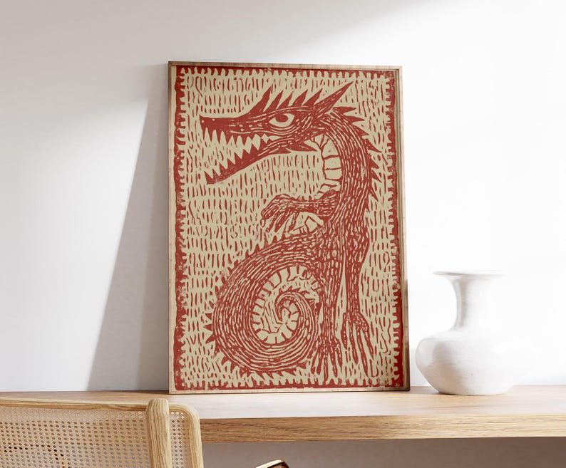 May include: Framed print featuring a red dragon illustration on a beige background. The dragon has sharp teeth and claws, with a coiled tail. The artwork is in a wooden frame and rests on a wooden shelf. A white vase is next to the print.