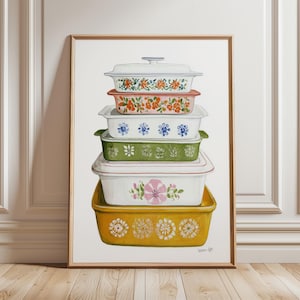 Op de afbeelding: Een ingelijste print met een stapel vintage ovenschalen. De schalen zijn in verschillende kleuren, waaronder geel, groen en wit, en versierd met bloemmotieven. Het kunstwerk wordt tentoongesteld op een houten vloer.