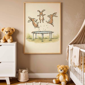 May include: A framed print featuring three brown rabbits jumping on a trampoline. The artwork has a light beige background with green grass and a blue trampoline. The frame is made of light wood. The room has a white dresser, a teddy bear, and a baby crib.