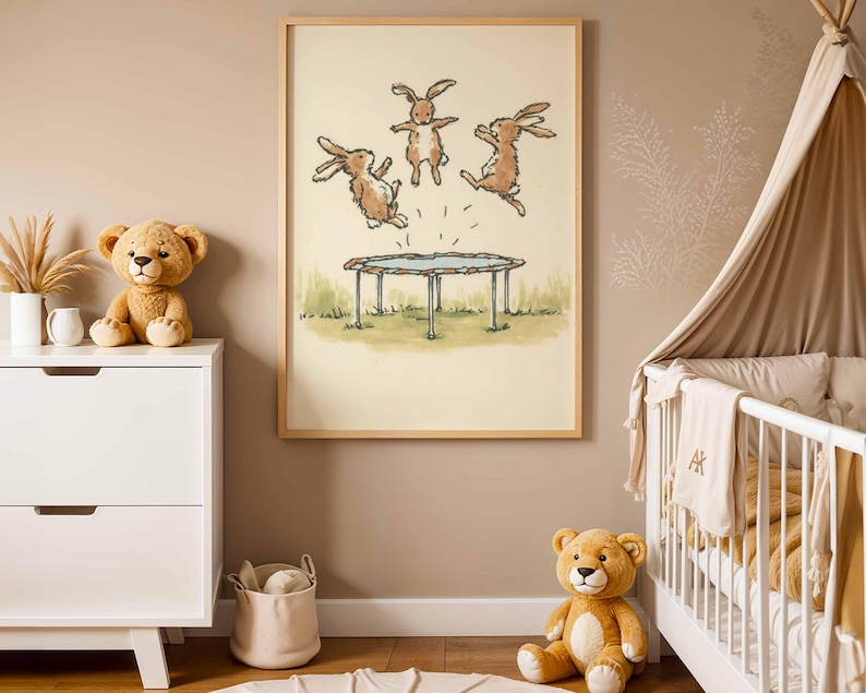 May include: A framed print featuring three brown rabbits jumping on a trampoline. The artwork has a light beige background with green grass and a blue trampoline. The frame is made of light wood. The room has a white dresser, a teddy bear, and a baby crib.