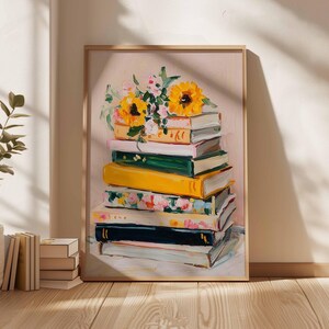 May include: A framed art print featuring a stack of books with a bouquet of sunflowers and other flowers on top. The books are painted in various colors, including yellow, green, and floral patterns. The background is a soft pink.