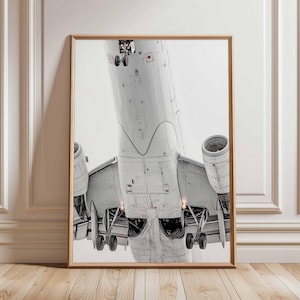 Könnte beinhalten: Gerahmter Druck eines weißen Flugzeugs von unten, mit Fahrwerk und Triebwerken deutlich sichtbar. Das Bild, aus einem niedrigen Winkel aufgenommen, ist vor einem hellen, bewölkten Himmel platziert.