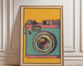 May include: A framed art print featuring a vintage camera illustration. The camera is teal and pink, with a large lens and a yellow background. The artwork is displayed in a wooden frame, leaning against a white wall and wooden floor.