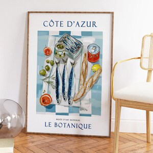 May include: A framed art print featuring a still life of food items. The image includes sardines, olives, bread, and citrus fruits on a blue and white checkered cloth. The text "C&Ocirc;TE D'AZUR" and "LE BOTANIQUE" are visible.
