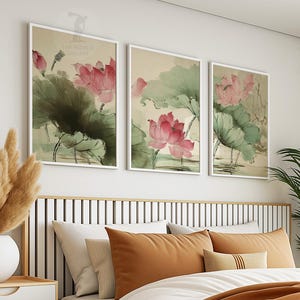 May include: Three framed art prints featuring watercolor paintings of pink lotus flowers and green leaves. The artwork is displayed above a bed with orange and white pillows. The prints are in white frames.