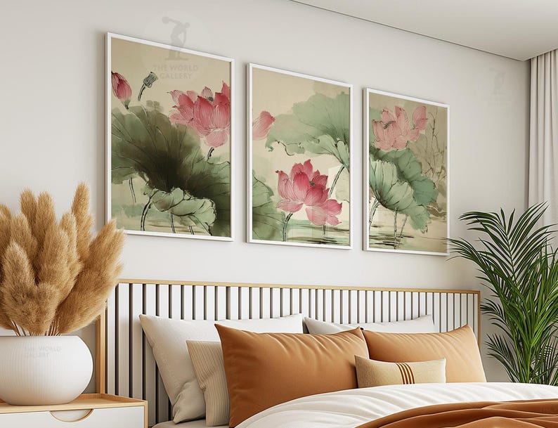May include: Three framed art prints featuring watercolor paintings of pink lotus flowers and green leaves. The artwork is displayed above a bed with orange and white pillows. The prints are in white frames.