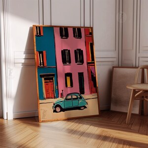 May include: A framed art print depicts a colorful street scene with a teal car. The buildings are painted in vibrant hues of blue, pink, and red, with black window shutters and orange doors. The artwork is leaning against a white wall and wooden floor.