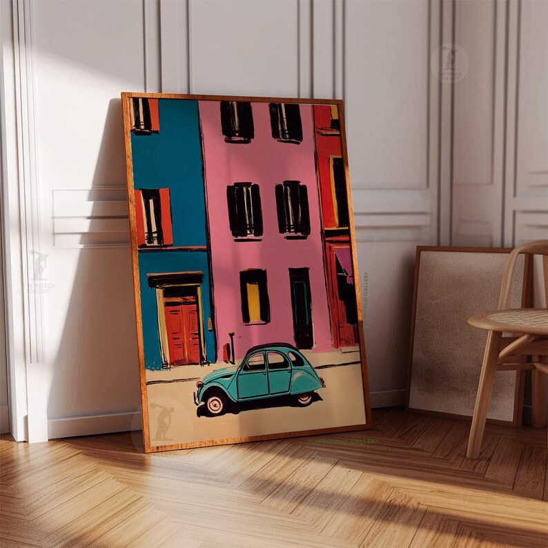 May include: A framed art print depicts a colorful street scene with a teal car. The buildings are painted in vibrant hues of blue, pink, and red, with black window shutters and orange doors. The artwork is leaning against a white wall and wooden floor.