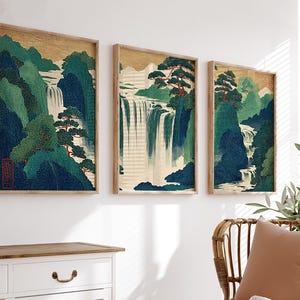 May include: Three framed art prints depicting a waterfall scene. The artwork features a gold background, green and blue mountains, and a cascading waterfall. The prints are in wooden frames and are displayed on a white wall.