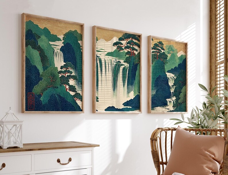 May include: Three framed art prints depicting a waterfall scene. The artwork features a gold background, green and blue mountains, and a cascading waterfall. The prints are in wooden frames and are displayed on a white wall.