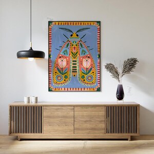 Pode incluir: Uma pintura colorida de arte popular de uma mariposa com padr&otilde;es intrincados e desenhos florais, emoldurada em preto. A obra de arte est&aacute; pendurada acima de um arm&aacute;rio de madeira com um vaso de flores secas.