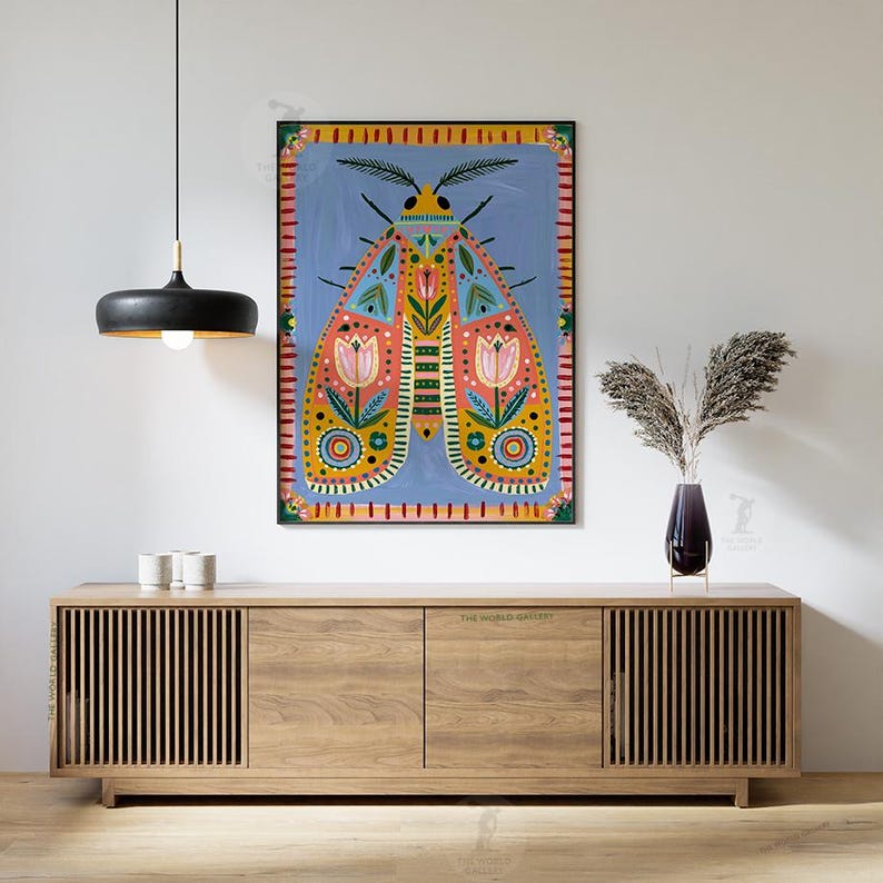 Pode incluir: Uma pintura colorida de arte popular de uma mariposa com padr&otilde;es intrincados e desenhos florais, emoldurada em preto. A obra de arte est&aacute; pendurada acima de um arm&aacute;rio de madeira com um vaso de flores secas.