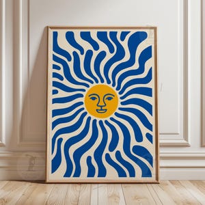 May include: A framed art print featuring a stylized sun design. The sun has a yellow face with blue wavy rays against a cream background. The artwork is in a wooden frame and is displayed on a white wall.
