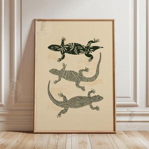 Peut inclure: Impression d'art encadrée représentant trois lézards stylisés en noir sur fond crème. Les lézards présentent des motifs, notamment floraux et géométriques. L'œuvre est dans un cadre en bois.