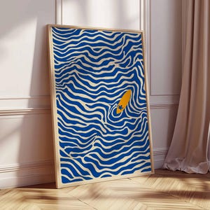 May include: Abstract art print featuring a blue and cream wavy pattern with an orange surfboard. The artwork is framed in a light wood frame and leans against a white wall and wooden floor. The art print adds a modern touch to any space.