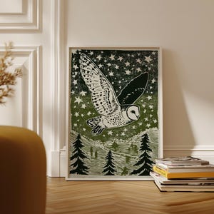 Op de afbeelding: Een ingelijste kunstdruk met een witte uil in vlucht tegen een donkergroene sterrenhemel. De uil heeft gedetailleerde vleugelpatronen. Hieronder is een boslandschap met bomen te zien. Het kunstwerk wordt op een houten vloer getoond.