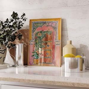 May include: A framed watercolor painting of a colorful door with a floral design. The door is set in a pink archway with a yellow background. The painting is on a white countertop with a vase, a pitcher, and a bowl of lemons.