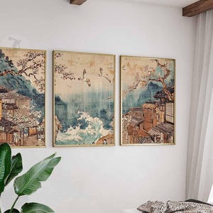 May include: A set of three framed prints depicting a Japanese landscape with traditional houses, cherry blossoms, and a view of the ocean. The prints are in a muted color palette with shades of blue, green, and brown.