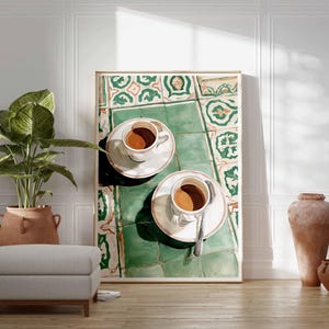 May include: A framed artwork depicts two cups of coffee on saucers, resting on a green and white tiled surface. The artwork is propped against a white wall, with a potted plant, ottoman, and decorative vases in the foreground.