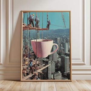 以下が含まれることがあります： シュールなコラージュデザインの額入りアートプリント。建設作業員が建物のフレームに描かれ、大きなピンク色のティーカップが都市景観に重ねられています。アートワークは、明るい茶色の木製フレームに額装されています。