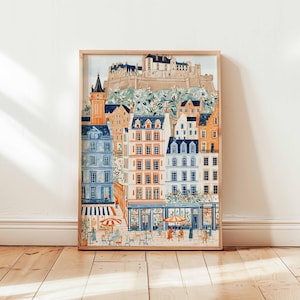 May include: A framed watercolour print of a European cityscape. The artwork depicts colourful buildings with orange, blue, and white facades, a castle on a hill, and outdoor cafes. The print is displayed on a wooden floor.