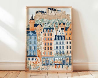 May include: A framed watercolor print of a European cityscape. The artwork depicts colorful buildings with orange, blue, and white facades, a castle on a hill, and outdoor cafes. The print is displayed on a wooden floor.