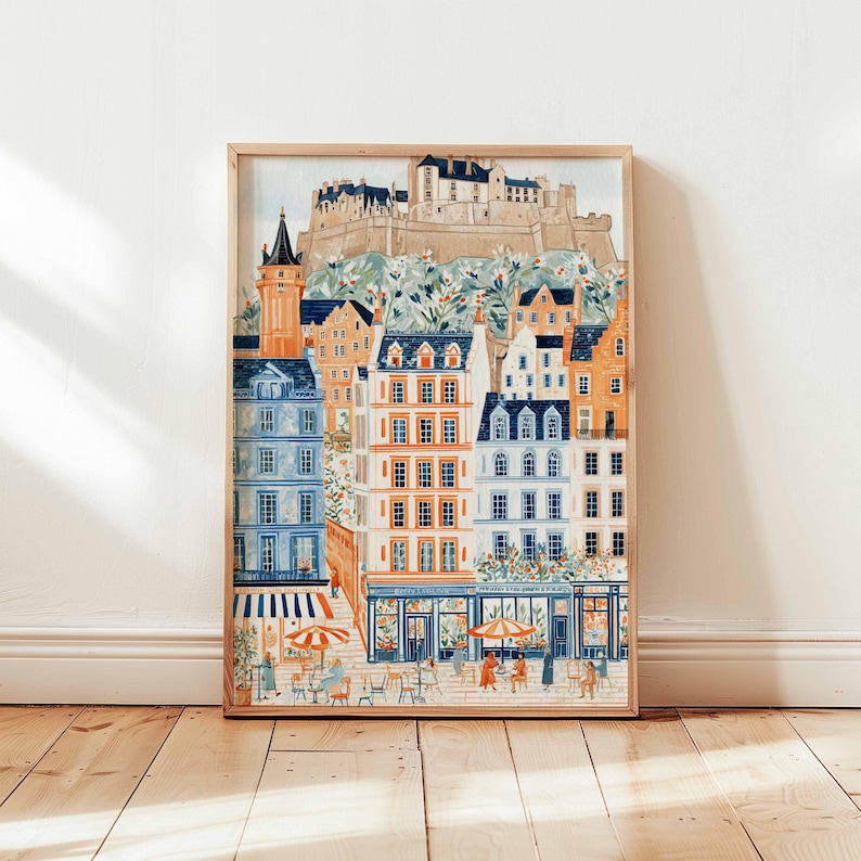 May include: A framed watercolour print of a European cityscape. The artwork depicts colourful buildings with orange, blue, and white facades, a castle on a hill, and outdoor cafes. The print is displayed on a wooden floor.