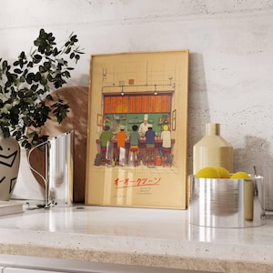 May include: A framed print depicting a Japanese ramen shop scene. The artwork features a chef behind a counter serving customers seated at stools. The print has Japanese text and is displayed on a countertop with decorative items.