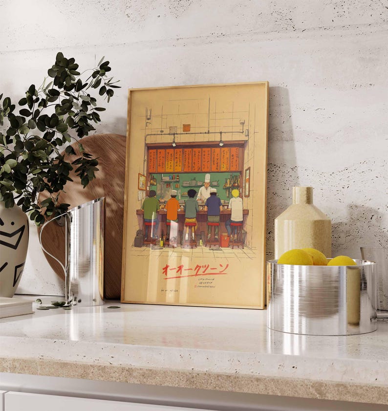 May include: A framed print depicting a Japanese ramen shop scene. The artwork features a chef behind a counter serving customers seated at stools. The print has Japanese text and is displayed on a countertop with decorative items.
