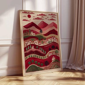 May include: A framed art print showing a stylized landscape. The artwork features a red sun, mountains, and rolling hills with white buildings and trees. The colour palette includes shades of red, pink, and green. The frame is a light wood colour.