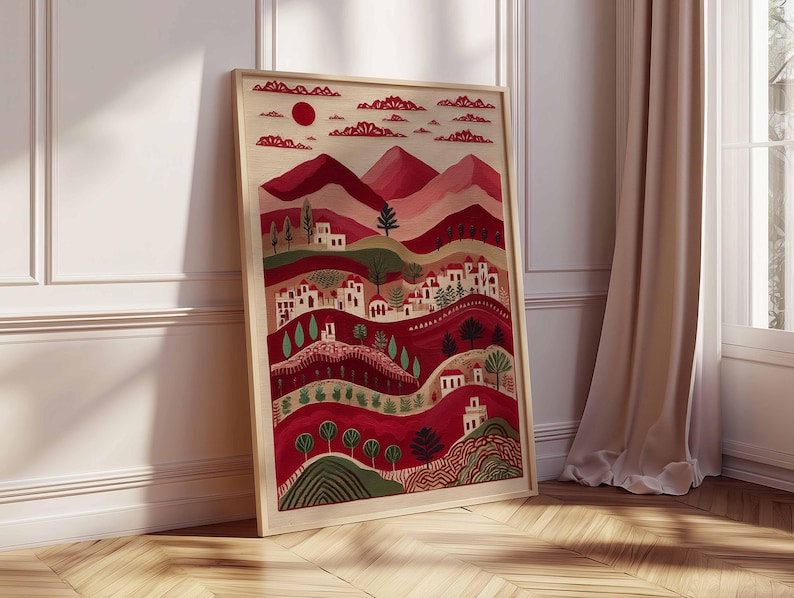 May include: A framed art print showing a stylized landscape. The artwork features a red sun, mountains, and rolling hills with white buildings and trees. The colour palette includes shades of red, pink, and green. The frame is a light wood colour.