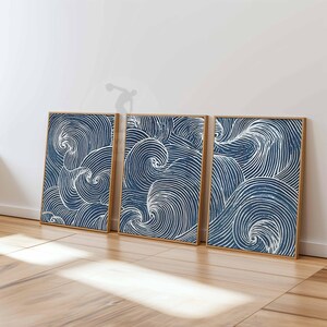 May include: Three framed art prints with a blue and white wave pattern. The artwork features swirling lines in a navy blue background, creating a dynamic and textured effect. The frames are a natural wood color, adding warmth to the overall aesthetic.