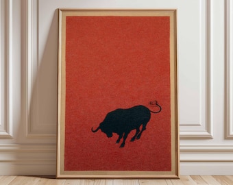 May include: Framed art print featuring a black silhouette of a bull on a textured red background. The artwork is encased in a light brown wooden frame, suitable for home or office decor. The bull is depicted in a dynamic, charging pose.