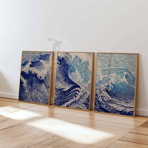 May include: Three framed prints featuring a stylized depiction of ocean waves in shades of blue and beige. The artwork has a woodblock print aesthetic, with detailed lines and textures. The frames are a light brown color.