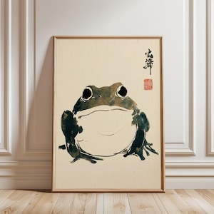 May include: A framed art print depicting a stylized frog illustration in black, green, and white. The frog has large, dark eyes and a wide mouth. Japanese characters and a red seal are in the upper right corner. The artwork is in a wooden frame.