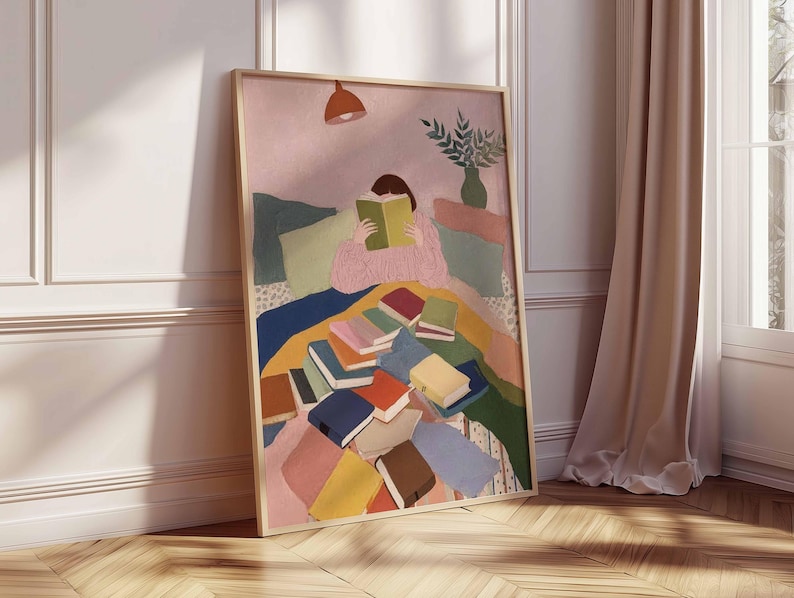 May include: A framed art print depicts a person reading amidst a pile of books. The artwork features a soft color palette with pink, blue, and yellow hues. A lamp hangs above the figure, and a vase of greenery sits in the background.