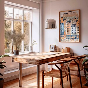 May include: A light-filled room featuring a wooden desk and two woven chairs. A framed print of a European town adorns the wall. A large window provides a view of trees. A silver pendant light hangs above the desk.